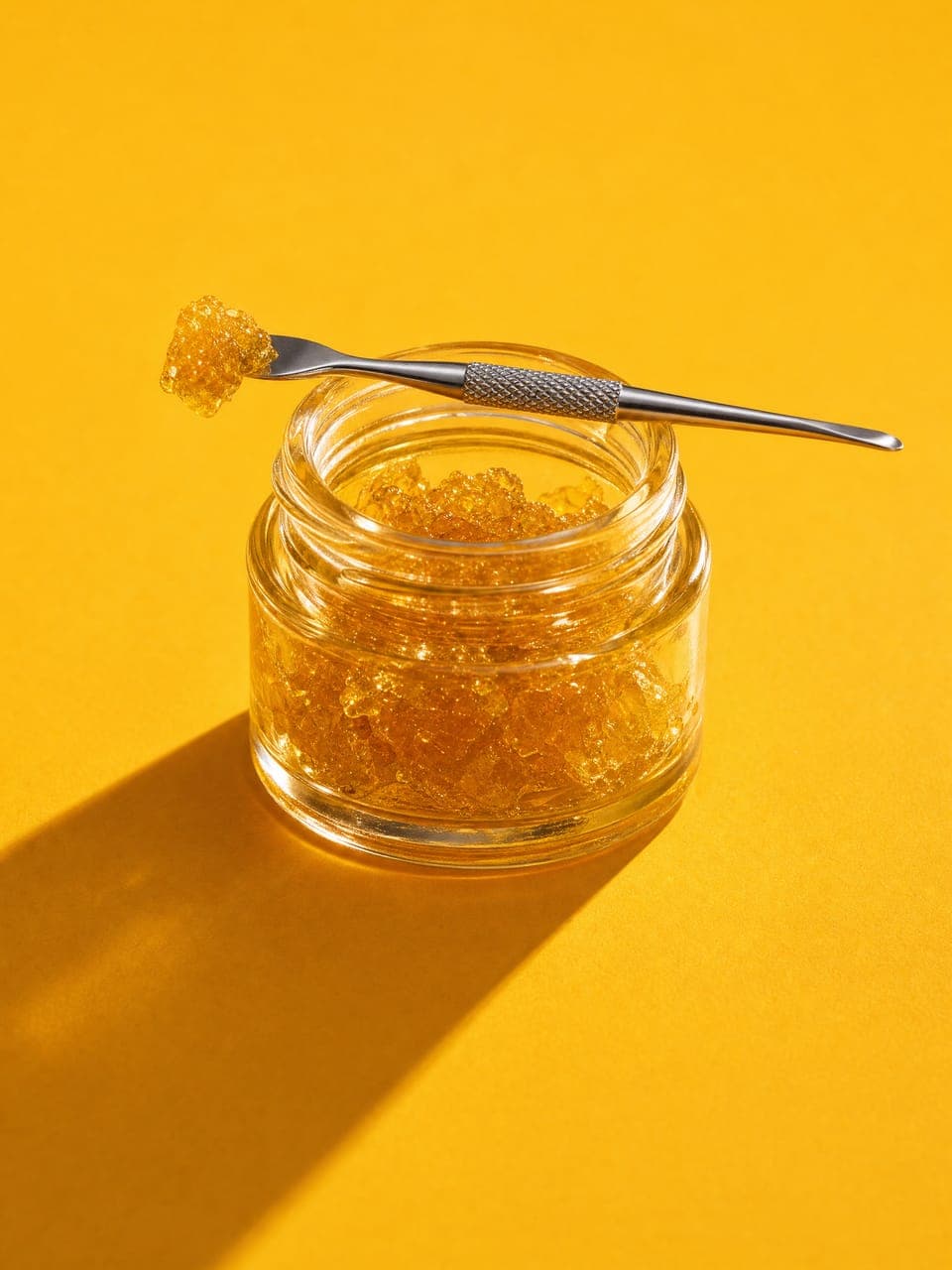 A glass jar of golden wax with a dab tool on a yellow backdrop