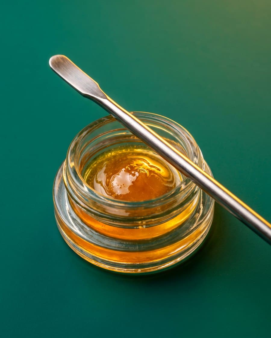 A glass jar of amber concentrate with a dab tool on a green backdrop